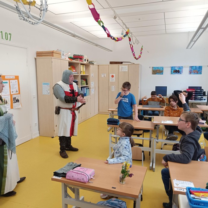 Schülerinnen und Schüler melden sich, um dem verkleideten Ritter fragen zu stellen (vergrößerte Bildansicht wird geöffnet)